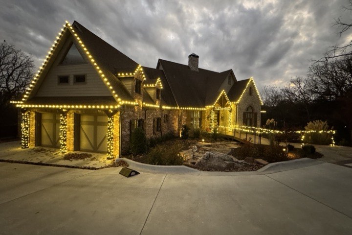 A stone and brick home at dusk with warm white Christmas lights along every roofline and around the garage