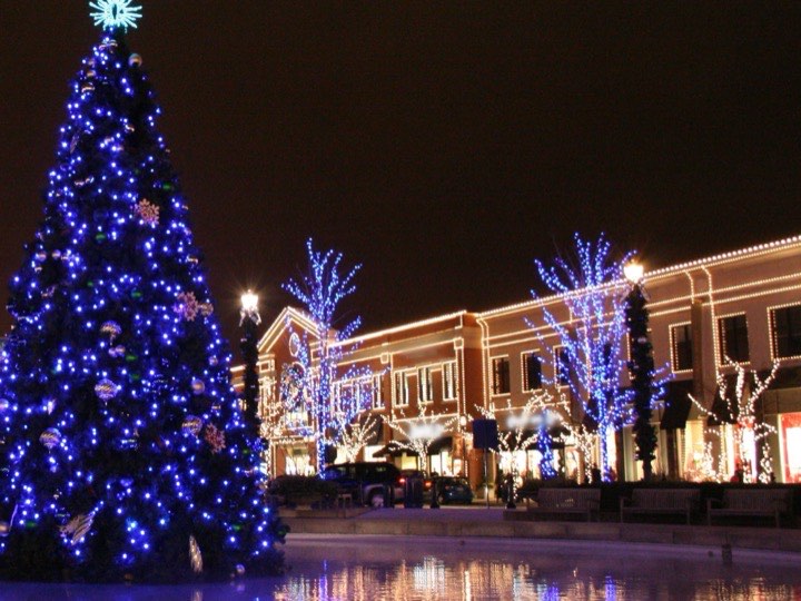 A shopping center at night with lights along the rooflines, light-wrapped trees and a lit Christmas tree